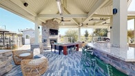 Located at 489 N McDowell Blvd in Petaluma, California, this thoughtfully designed outdoor amenity space offers a variety of recreational and social environments within a well-appointed community setting. At the heart of the space is a covered pavilion featuring a soaring vaulted ceiling with exposed white-painted beams, ceiling fans, and radiant heaters for year-round comfort. A floor-to-ceiling stacked stone fireplace with a mounted flat-screen TV serves as the focal point, flanked by a shuffleboard game table and a stone-faced outdoor bar with counter-height seating. The pavilion floor is finished with a decorative paver pattern, adding texture and visual interest throughout. Adjacent to the pavilion, a circular spa offers a relaxing retreat, surrounded by a paver deck and lounge seating with open views of the surrounding landscape. A manicured lawn area provides open green space, bordered by glass panel fencing, landscape plantings, and pathway lighting. An additional open-air patio features dining tables shaded by large market umbrellas, creating a comfortable space for outdoor gatherings. The multi-building residential backdrop, combined with mature trees and a clear California sky, contributes to the inviting and community-centered ambiance of this outdoor space.