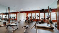 This fitness center is designed to support a comprehensive workout experience within a single, open-plan space. The room is immediately distinguished by its bold red accent walls adorned with diamond-shaped geometric patterns in gold and tan, lending the space an energetic and visually striking atmosphere. Durable rubber flooring covers the entire area, providing traction and cushioning suited for a variety of exercise activities.

The cardio section is well-appointed, featuring multiple treadmills, elliptical trainers, and stationary bikes positioned along a wall of windows. These windows allow ample natural light to fill the room and offer views of the surrounding outdoor greenery, creating a more open and inviting environment during workouts. A wall-mounted flat-screen television is positioned for easy viewing from the cardio equipment.

Floor-to-ceiling mirrors framed in dark wood line one wall, visually expanding the space and allowing users to monitor their form. The strength training area includes a multi-function cable machine, a weight rack with dumbbells, a rowing machine, a flat bench, and suspension training straps. Recessed ceiling lighting supplements the natural light, ensuring consistent illumination throughout. A separate door provides access to an adjacent area, adding functional convenience to the overall layout.