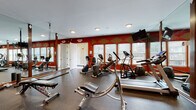 This fitness center is designed to support a comprehensive workout experience within a single, open-plan space. The room is immediately distinguished by its bold red accent walls adorned with diamond-shaped geometric patterns in gold and tan, lending the space an energetic and visually striking atmosphere. Durable rubber flooring covers the entire area, providing traction and cushioning suited for a variety of exercise activities.

The cardio section is well-appointed, featuring multiple treadmills, elliptical trainers, and stationary bikes positioned along a wall of windows. These windows allow ample natural light to fill the room and offer views of the surrounding outdoor greenery, creating a more open and inviting environment during workouts. A wall-mounted flat-screen television is positioned for easy viewing from the cardio equipment.

Floor-to-ceiling mirrors framed in dark wood line one wall, visually expanding the space and allowing users to monitor their form. The strength training area includes a multi-function cable machine, a weight rack with dumbbells, a rowing machine, a flat bench, and suspension training straps. Recessed ceiling lighting supplements the natural light, ensuring consistent illumination throughout. A separate door provides access to an adjacent area, adding functional convenience to the overall layout.