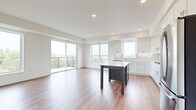 This thoughtfully designed two-bedroom, one-bathroom residence offers a bright and contemporary living experience across a single floor. The open-concept kitchen and living area serves as the heart of the home, featuring warm hardwood-style flooring, recessed lighting, and large windows that flood the space with natural light. Sliding glass doors lead to a private balcony, offering pleasant outdoor views of surrounding greenery.

The kitchen is well-appointed with crisp white shaker-style cabinetry, light quartz countertops, a gray subway tile backsplash, a deep stainless steel undermount sink, and a full suite of stainless steel appliances including a gas cooktop, double wall oven, refrigerator, and dishwasher. A central island with a contrasting dark base provides additional workspace and seating. The living room is anchored by a sleek fireplace with a wood-toned mantel and built-in shelving.

Both bedrooms are carpeted and finished in soft neutral tones. The spacious primary bedroom includes a walk-in closet with built-in shelving. A second bedroom features a tall vertical window with treetop views and additional built-in shelving. The full bathroom offers a tub-shower combination with white subway tile surround. A dedicated laundry room includes a full-size front-loading washer and dryer.