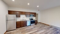 This single-floor unit offers a clean, modern aesthetic throughout, with consistent wood-look flooring and neutral white walls that create a bright, cohesive atmosphere across all rooms. The open-concept kitchen is a standout feature, outfitted with rich dark-stained cabinetry extending to the upper walls, light stone-look countertops, a double-basin sink, and a full suite of stainless steel appliances including a refrigerator, dishwasher, electric range, and over-the-range microwave. The kitchen flows naturally into a generously sized living area, which benefits from recessed lighting and baseboard heating along the perimeter walls, lending the space both functionality and comfort. A dedicated laundry room provides washer and dryer hookups, offering added convenience. The bedrooms are well-proportioned, each featuring the same wood-look flooring, clean white walls, and ample overhead clearance that contributes to an open, airy feel. The full bathroom is finished with a walk-in shower accented by neutral tile, a dark wood vanity with a white countertop, a large mirror, and wood-look flooring that ties the space together. Throughout the unit, warm wood-toned baseboard trim adds a grounding detail that complements the overall modern and functional design.