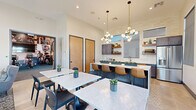 This contemporary single-floor community amenity space is thoughtfully designed to foster social interaction and everyday comfort. The open-plan layout seamlessly connects a fully equipped kitchen, casual dining areas, and a relaxed lounge, all bathed in abundant natural light from large windows and sliding glass doors.

The kitchen is a standout feature, outfitted with slate-blue cabinetry, a white quartz countertop island with seating for several bar-height leather chairs, a gleaming subway tile backsplash, open walnut floating shelves, brushed gold fixtures, and stainless steel appliances. The space is illuminated by striking globe pendant chandeliers suspended from elevated ceilings, adding a refined, modern touch.

The dining area offers multiple marble-top tables paired with sleek black chairs, accented by sculptural wall art and dramatic pendant lighting. A lounge area features a large wall-mounted television, navy cabinetry with gold hardware, and comfortable seating that opens directly onto a sunny outdoor patio through floor-to-ceiling sliding glass doors.

The fitness center, accessible via glass barn-style doors, is equipped with cardio and strength training machines, oversized industrial ceiling fans, and motivational wall murals, creating an energetic and functional workout environment. Throughout the space, light tile flooring, high ceilings with clerestory windows, and a consistent modern-transitional design palette unify every area cohesively.