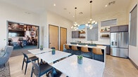 This contemporary single-floor community amenity space is thoughtfully designed to foster social interaction and everyday comfort. The open-plan layout seamlessly connects a fully equipped kitchen, casual dining areas, and a relaxed lounge, all bathed in abundant natural light from large windows and sliding glass doors.

The kitchen is a standout feature, outfitted with slate-blue cabinetry, a white quartz countertop island with seating for several bar-height leather chairs, a gleaming subway tile backsplash, open walnut floating shelves, brushed gold fixtures, and stainless steel appliances. The space is illuminated by striking globe pendant chandeliers suspended from elevated ceilings, adding a refined, modern touch.

The dining area offers multiple marble-top tables paired with sleek black chairs, accented by sculptural wall art and dramatic pendant lighting. A lounge area features a large wall-mounted television, navy cabinetry with gold hardware, and comfortable seating that opens directly onto a sunny outdoor patio through floor-to-ceiling sliding glass doors.

The fitness center, accessible via glass barn-style doors, is equipped with cardio and strength training machines, oversized industrial ceiling fans, and motivational wall murals, creating an energetic and functional workout environment. Throughout the space, light tile flooring, high ceilings with clerestory windows, and a consistent modern-transitional design palette unify every area cohesively.