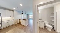 This well-designed single-floor residence offers a clean, contemporary aesthetic with a thoughtful open-concept layout. The kitchen serves as the centerpiece of the living area, featuring white shaker-style cabinetry that extends to the ceiling, stainless steel appliances including a refrigerator, electric range, and microwave, and a quartz-topped center island with an undermount sink and pendant lighting overhead. A subway tile backsplash and under-cabinet lighting add refined detail to the space. Light wood-look plank flooring flows throughout the main living areas, contributing to a bright and cohesive atmosphere. The living area receives ample natural light through large windows fitted with horizontal blinds, with views of the surrounding exterior. The bedroom features plush carpeting and a generously sized window that further enhances the sense of openness. Two bathrooms are present in the unit — one featuring a walk-in glass-enclosed shower, white shaker vanity, and built-in shelving, and a second, larger bathroom with an extended quartz vanity, wide wall mirror, and a tiled walk-in shower with a frameless glass door. Neutral wall tones and consistent finishes throughout create a cohesive and move-in-ready environment.