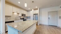 Located at 10491 Gandy Blvd N in Saint Petersburg, Florida, this single-floor residence showcases a clean, contemporary design with a bright and airy aesthetic throughout. The open-concept layout seamlessly connects the kitchen and living area, creating a spacious, flowing environment ideal for everyday living. The kitchen is a standout feature, equipped with white shaker-style cabinetry, a subway tile backsplash, granite countertops, a large center island with pendant lighting, and a full suite of stainless steel appliances. Wood-look plank flooring runs throughout the main living areas, complementing the neutral color palette and recessed lighting overhead. The living space benefits from natural light through a sliding glass door that opens to an outdoor balcony area with views of lush Florida greenery. The primary bathroom features a dual-sink vanity with granite countertops, white cabinetry, a glass-enclosed shower, and direct access to a spacious walk-in closet outfitted with wire shelving. A secondary bedroom is finished with plush carpeting and a paneled door. A dedicated laundry room houses a full-size washer and dryer with overhead wire shelving for added storage. The overall ambiance is modern, clean, and move-in ready.