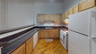 Located at 700 Stockton St in Richmond, Virginia, this single-floor loft-style unit showcases a compelling blend of industrial character and practical living. Throughout the space, warm-toned original hardwood floors run continuously, lending a sense of cohesion and rustic charm to every room.

The open main living area is defined by striking exposed beam ceilings with visible ductwork and piping, evoking classic industrial loft aesthetics. Large steel-framed windows allow natural light to filter in, illuminating the polished hardwood floors and the clean white walls. Industrial-style wall-mounted sconce lighting runs along a long hallway, reinforcing the utilitarian design sensibility.

The kitchen is well-appointed with light wood cabinetry, dark granite-style countertops, a full suite of white appliances including a refrigerator, electric range, and dishwasher, and a peninsula that opens toward the living area. The layout offers generous counter space and storage.

The bathroom features clean white tile flooring, a pedestal sink, a full bathtub with shower, and a vibrant mosaic-tiled shower curtain that adds a pop of color to the otherwise neutral palette.

Additional rooms with white walls and original hardwood floors provide flexible spaces suitable for use as bedrooms or offices, each with overhead lighting and small upper windows that bring in supplemental natural light.
