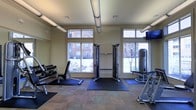 This single-floor fitness center is thoughtfully designed to support a comprehensive workout experience. The space is brightened by an abundance of natural light streaming through multiple large windows, offering views of a lush outdoor courtyard and neighboring brick buildings. Exposed ductwork runs along the neutral-toned ceiling, lending the room an open, industrial character.

The strength training area is well-appointed with a multi-functional cable machine positioned centrally, providing versatile resistance training options. To one side, a full dumbbell rack stocked with a range of weights sits alongside an adjustable workout bench, ideal for a variety of free weight exercises. On the opposite side, an additional strength machine offers targeted muscle training. A mounted flat-screen television is positioned in the upper corner, allowing users to stay entertained during workouts.

Protective rubber floor mats are strategically placed beneath the equipment, ensuring safety and floor preservation. The carpeted flooring throughout the rest of the space adds warmth and comfort. The overall ambiance is clean, functional, and inviting, making it a practical and motivating environment for residents looking to maintain an active lifestyle.