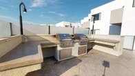 This rooftop terrace offers a well-appointed outdoor living experience designed for entertaining and relaxation. The centerpiece of the space is a built-in outdoor kitchen, constructed with a durable stucco-clad base and stone countertops in an L-shaped configuration. It features two stainless steel built-in grills side by side, each accompanied by stainless steel access doors for storage below — making it a fully functional setup for large-scale outdoor cooking. The tiled flooring throughout the terrace provides a clean, cohesive surface underfoot. A dedicated seating and dining area is positioned nearby, furnished with a round patio table, blue sling chairs, and a large red shade umbrella, creating a comfortable spot to dine al fresco. Curved raised planters are integrated into the terrace design, filled with low-maintenance succulents and ornamental plants that add a touch of greenery. A secondary, more intimate seating alcove offers additional lounging space with open views of the surrounding skyline. The perimeter is enclosed with stucco privacy walls and glass panel sections, maintaining an open feel while providing seclusion. The terrace benefits from abundant natural light throughout the day, making it an inviting outdoor retreat.