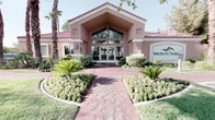This single-story community clubhouse at Rancho Del offers an inviting and thoughtfully designed gathering space that blends architectural elegance with a bright, resort-inspired aesthetic. The exterior makes a strong first impression with its Mediterranean-influenced façade, featuring a stucco finish, a red tile roof, and a covered entryway framed by tall windows. A decorative stone pathway winds through manicured landscaping, including tropical palms and lush greenery, leading to the glass double-door entrance.

Inside, the main gathering area is defined by dramatic vaulted ceilings with exposed white-painted beams that rise to a striking apex, flooding the space with a sense of openness and grandeur. Natural light pours in through a large gable-end window and glass doors at the far wall, offering views of the surrounding outdoor greenery. The interior features warm hardwood flooring throughout, anchored by a stone-clad fireplace with a built-in media unit that serves as a focal point on one end of the room. Comfortable seating arrangements in teal and aqua tones are thoughtfully arranged across the space, creating distinct lounge areas suited for relaxation and socializing. Recessed lighting and decorative fixtures complement the natural light, completing the bright and airy ambiance of this well-appointed community space.