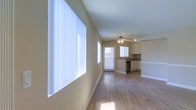 This single-floor space offers a clean, open-concept layout that connects the living area and kitchen in a cohesive and functional flow. The living area is immediately welcoming, characterized by large windows dressed with vertical blinds that allow abundant natural light to pour in, illuminating the warm, hardwood-style flooring that extends throughout the space. Neutral beige walls provide a versatile backdrop, keeping the atmosphere bright and airy.

The kitchen sits at the far end of the open layout and is well-appointed with light-toned cabinetry, a countertop workspace, and a built-in dishwasher, making it both practical and visually unobtrusive. A secondary window above the kitchen sink and a glass-panel door further enhance the natural light, creating a sense of openness that carries through the entire floor plan.

A ceiling fan with an integrated light fixture is centrally positioned overhead, providing both air circulation and supplemental lighting. Crisp white trim and baseboards add a polished finishing detail throughout. The overall ambiance is clean, minimal, and move-in ready, offering a versatile canvas suitable for a variety of interior styles and furnishing arrangements.