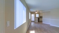 This single-floor space offers a clean, open-concept layout that connects the living area and kitchen in a cohesive and functional flow. The living area is immediately welcoming, characterized by large windows dressed with vertical blinds that allow abundant natural light to pour in, illuminating the warm, hardwood-style flooring that extends throughout the space. Neutral beige walls provide a versatile backdrop, keeping the atmosphere bright and airy.

The kitchen sits at the far end of the open layout and is well-appointed with light-toned cabinetry, a countertop workspace, and a built-in dishwasher, making it both practical and visually unobtrusive. A secondary window above the kitchen sink and a glass-panel door further enhance the natural light, creating a sense of openness that carries through the entire floor plan.

A ceiling fan with an integrated light fixture is centrally positioned overhead, providing both air circulation and supplemental lighting. Crisp white trim and baseboards add a polished finishing detail throughout. The overall ambiance is clean, minimal, and move-in ready, offering a versatile canvas suitable for a variety of interior styles and furnishing arrangements.