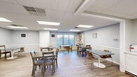 Located at 15075 Lincoln St in Oak Park, Michigan, this single-floor community room offers a well-designed, open-concept layout thoughtfully arranged to accommodate group gatherings and collaborative activities. The main area features light wood-look flooring, soft blue-gray walls with white wainscoting, and a drop ceiling fitted with multiple large recessed panel lights that provide consistent, even illumination throughout the space. Several dining-style tables with mixed seating — including traditional chairs and modern tulip-style seats — are arranged to maximize flexibility for various group configurations. A large window dressed with vertical blinds along one wall invites natural light and provides views of the outdoors, adding to the room's open and welcoming atmosphere. Motivational wall signage reading "believe," "inspire," and "stay awhile" contributes to a warm, encouraging ambiance. Built-in white shelving units offer practical display and storage space, while a compact kitchenette with white cabinetry and a sink provides added convenience. A wall-mounted television is also present, supporting presentations or media use. The space includes a full bathroom with a white vanity, large mirror, and wall-mounted soap dispensers, ensuring complete functionality for extended gatherings.