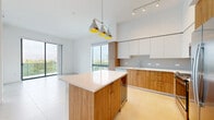 Located on the penthouse level at 999 NW Seventh St in Miami, FL, this 3-bedroom, 2-bathroom residence offers a thoughtfully designed layout with modern finishes throughout. The open-concept kitchen is a standout feature, showcasing warm wood cabinetry paired with crisp white upper cabinets, quartz countertops, a large center island with built-in wine cooler, stainless steel appliances, and a tiled backsplash. A trio of contemporary pendant lights with gold-toned interiors adds a stylish focal point above the island. Light porcelain tile flooring runs throughout, contributing to a clean, cohesive aesthetic. The bedrooms are bright and airy, with large floor-to-ceiling windows and sliding glass doors that invite abundant natural light and offer views of the surrounding cityscape. A modern barn-style sliding door provides a functional and design-forward detail in one of the bedrooms. The primary bathroom features a double-sink vanity with wood cabinetry, a white quartz countertop, and a walk-in glass shower, while the secondary bathroom includes a full soaking tub with tiled surround. The penthouse level also provides access to an outdoor terrace with sweeping panoramic views of the Miami skyline and lush green canopy below.