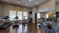 This dedicated fitness center is thoughtfully designed to support a comprehensive workout experience. The space features rich hardwood flooring throughout, lending a polished and durable surface well-suited for both cardio and strength training activities. Elevated ceilings with recessed lighting create an open, airy atmosphere, while multiple large windows dressed with white blinds allow generous natural light to fill the room and offer views of surrounding greenery.

The cardio section is equipped with treadmills, an elliptical trainer, and a stationary recumbent bike, all positioned near the windowed wall to make workouts more enjoyable. A wall-mounted flat-screen television is conveniently placed for entertainment during exercise sessions.

The strength training area is well-appointed with a variety of cable machines, a multi-functional weight bench, resistance equipment, and a rack of dumbbells in varying sizes. Exercise balls in multiple sizes add versatility for functional fitness routines. Floor-to-ceiling mirrors line one wall, enhancing the sense of space while allowing users to monitor their form during workouts.

The fitness center is accessed through elegant white-framed glass double doors, contributing to its clean, upscale aesthetic. The neutral gray walls and white trim provide a calm, focused environment conducive to exercise. Overall, this fitness center combines practical functionality with a refined, well-maintained design.