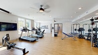 This generously sized fitness center offers a comprehensive workout environment across a single open floor plan. The space features light hardwood-style flooring, white walls, and recessed lighting complemented by ceiling fans, creating a bright and comfortable atmosphere for exercise.

The main workout area is thoughtfully organized to accommodate a wide range of fitness activities. A dedicated cardio zone houses multiple treadmills and elliptical machines, while the strength training section includes a power rack with weight bench, cable machines, selectorized resistance equipment from True Fitness, a dumbbell rack, and plyo boxes for functional training. Motivational phrases such as "Make It Happen" and "Passion" are displayed prominently on the walls, adding an energizing ambiance to the space.

Natural light flows in through large windows along the perimeter, offering views of the surrounding outdoor area. Structural columns are integrated throughout the floor plan, subtly defining zones without interrupting the open layout.

A separate studio room features a serene nature-inspired wall mural depicting tall grasses, warm-toned hardwood flooring, a ceiling fan, and a wall-mounted interactive fitness display — ideal for yoga, stretching, or guided workout sessions. Together, these spaces form a well-rounded, modern fitness facility designed to support a variety of exercise routines and wellness goals.