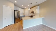 This single-floor space offers a clean, contemporary design throughout. The open living area features light wood-look flooring, neutral warm-toned walls, and a ceiling fan overhead. A full-glass sliding door leads to a private balcony with metal railing, allowing natural light to fill the space and providing views of the surrounding community.

The kitchen is a standout feature, designed with two-tone cabinetry — white upper cabinets paired with muted blue-gray lower cabinets — and light quartz countertops. A peninsula with an integrated sink and pendant lighting above serves as a functional focal point. Stainless steel appliances, including a gas range, built-in microwave, and refrigerator, are complemented by a white subway tile backsplash.

The bedroom continues the neutral palette with plush carpeting, a ceiling fan, a roller shade window, and a paneled white door. The bathroom is finished with light wood-look tile flooring, a tiled tub-shower combo with a curved glass door, a blue-gray vanity with chrome hardware, a white quartz countertop, and an illuminated frameless mirror, creating a polished and functional space.