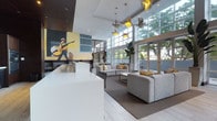 This single-floor space presents a sleek, contemporary lobby or communal lounge that blends artistic character with modern design. The interior is anchored by soaring double-height ceilings that flood the space with natural light through an expansive floor-to-ceiling grid window wall, offering views of lush greenery beyond. A striking large-scale mural of a musician playing guitar dominates one wall, injecting a vibrant cultural energy and artistic identity into the environment. Overhead, a sculptural pendant lighting installation featuring bird-shaped fixtures in gold and silver tones adds a whimsical yet sophisticated focal point. The seating area is thoughtfully arranged with plush, low-profile grey sofas and accent pillows in warm mustard yellow, creating an inviting and cohesive color palette. Light-toned tile flooring and a clean white reception counter reinforce the bright, open atmosphere. Direct access to an outdoor terrace or courtyard is visible through glass-paneled doors, further enhancing the seamless indoor-outdoor connection. The overall ambiance is one of stylish comfort — a well-curated space that balances bold artistic expression with functional, communal hospitality design.