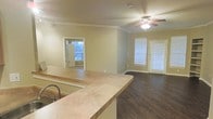 Located at Settler's Gate Apartments in Allen, Texas, this single-floor residence offers a well-designed open-concept layout that seamlessly connects the kitchen and living areas. The kitchen features a curved breakfast bar counter with an undermount sink, providing a natural division between the cooking and living spaces while maintaining an open and airy feel. Dark hardwood-style flooring runs throughout the main living area, adding warmth and a touch of elegance to the space. Crown molding accents the ceilings, lending a refined architectural detail that elevates the overall aesthetic. A ceiling fan with a light fixture is centrally positioned in the living area, ensuring comfortable airflow and ambient lighting. Multiple windows dressed with blinds invite generous natural light into the space, creating a bright and welcoming atmosphere. A glass-panel exterior door provides additional light and convenient access to the outdoors. An adjacent room, visible through a doorway, features a window that further enhances the natural brightness of the unit. The neutral wall tones throughout complement the flooring and trim, resulting in a cohesive and move-in-ready interior that offers a comfortable and functional living environment.