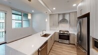 This two-floor residence showcases a clean, contemporary design throughout, with wood-look flooring and crisp white walls creating a cohesive, modern aesthetic. The main level centers around a well-appointed kitchen featuring light wood cabinetry, quartz countertops with subtle veining, and a peninsula with an integrated sink and dishwasher. Stainless steel appliances, including a gas range with a stainless steel hood and a French door refrigerator, are complemented by a marble-look tile backsplash. Recessed lighting and pendant fixtures illuminate the space, while a window allows natural light to filter in. A staircase with dark wood treads and a black metal railing connects the two levels, anchoring the open living area below.

The upper level houses the bedrooms, each offering light-toned wood-look flooring, neutral walls, and windows with roller shades that provide views of the surrounding neighborhood. Two bathrooms serve the residence: one features a frameless glass walk-in shower with floor-to-ceiling tile and a quartz vanity, while the second offers a soaking tub with a sliding glass enclosure, tiled surround, and a matching quartz vanity. A smart thermostat is also visible, adding a layer of modern convenience throughout the home.