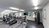 This fitness center offers a practical and well-organized space for a full workout experience. The room is lined with large mirrors along one wall, enhancing the sense of space and allowing users to monitor their form during exercise. A row of True-brand treadmills and elliptical machines takes center stage, providing ample cardio options for users of varying fitness levels. Complementing the cardio equipment is a cable resistance training machine positioned near the far wall, offering versatile strength training capabilities. A wall-mounted flat-screen television is centrally placed, providing entertainment during workouts, while informational fitness posters are displayed nearby for reference. Natural light filters in through multiple windows fitted with blinds, creating a bright and inviting atmosphere. The flooring features a dark, durable surface well-suited to high-traffic exercise use, and the textured ceiling houses rows of fluorescent lighting that ensure the room is evenly and thoroughly illuminated. The overall layout is efficient and functional, making the most of the available space to accommodate a range of fitness activities in a clean, focused environment.