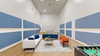 Located at 1700 Old England Loop in Sanford, Florida, this single-floor amenity space serves as a dynamic social and recreational hub. The room immediately impresses with its dramatically high ceilings, which lend an airy, open feel and amplify the sense of spaciousness throughout. Warm-toned hardwood-style flooring runs the length of the space, providing a cohesive and inviting foundation.

The lounge area is thoughtfully furnished with a mix of seating options, including a white sofa accented with blue throw pillows, a blue sectional, and a vibrant orange accent chair, all arranged around a gold-framed glass coffee table atop a patterned area rug. A bold design choice defines the walls: large blue acoustic panels are mounted in a grid-like arrangement, serving both a functional sound-dampening purpose and a striking visual element. Decorative triangle wall art adds a playful, geometric layer to the overall aesthetic.

The game zone features a billiards table and a foosball table, making the space well-suited for casual recreation and social gatherings. Large pass-through windows and a central doorway provide a direct sightline into an adjacent, fully equipped fitness center, seamlessly connecting active and leisure amenities. Recessed lighting throughout ensures the space remains well-lit and welcoming at all times.