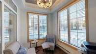 This single-floor space is thoughtfully designed to balance comfort and practicality across several distinct areas. The entry vestibule welcomes visitors with large glass panels and transom windows that allow natural light to fill the space, complemented by tile flooring and wall-mounted security and fire safety systems alongside a keyed interior door for controlled access.

Adjacent to the entry, a cozy lounge area serves as the centerpiece of the space. It features a coffered ceiling with a decorative chandelier, warm wood-trimmed windows dressed with horizontal blinds, and abundant natural light with outdoor views. Seating includes two upholstered accent chairs with patterned throw pillows arranged around small side tables, creating an inviting and functional sitting area. Baseboard heating runs along the exterior walls, and the rich tile flooring continues throughout.

A designated wifi room, accessed through a wood-framed glass door etched with signage, provides a separate functional zone equipped with a printer station, a rustic industrial-style console table, and additional seating. Built-in cabinetry with granite countertops is visible beyond the doorway, suggesting a kitchenette or service counter nearby. The cohesive use of warm wood tones, neutral walls, and tile flooring throughout gives the space a unified, welcoming aesthetic.