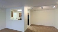 This single-floor space showcases an open-concept layout that connects the living area to the kitchen through a functional pass-through window, enhancing the sense of openness between the two areas. The living space is finished with wall-to-wall carpeting in a neutral tone, providing a comfortable and understated foundation. Crisp white walls throughout contribute to a clean, bright atmosphere.

The kitchen is partially visible through the pass-through opening and features medium-toned wood cabinetry, a black refrigerator, and a black microwave, offering a cohesive, practical aesthetic. A sink is positioned at the pass-through counter, allowing for convenient interaction between the kitchen and living areas.

Overhead, a track lighting system with adjustable spotlights runs along the ceiling of the main living space, offering flexible and focused illumination. A textured ceiling adds a subtle layer of visual detail throughout the unit. A skylight or recessed light panel in the kitchen provides additional brightness to that workspace. Neutral-toned baseboards frame the floors, completing the clean and functional design of this well-organized single-level space.