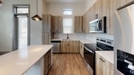 This single-floor residence at 3508 Mount Moriah Rd in Durham, North Carolina, presents a contemporary design with clean lines, neutral tones, and quality finishes throughout. The open-concept kitchen serves as a central highlight, featuring wood-grain cabinetry with matte black hardware, crisp white quartz countertops, a large center island, and a full suite of stainless steel appliances including a refrigerator, range, microwave, and dishwasher. Striking pendant lights hang above the island, complementing elevated ceilings that lend the kitchen and living areas a notably airy, spacious feel. Natural light filters in through strategically placed windows, including transom-style windows positioned high on the walls.

Wood-look plank flooring flows through the main living and kitchen areas, while plush carpet covers the bedrooms, adding warmth and comfort. A wide hallway connects the living spaces to the private quarters, with a walk-in closet featuring wire shelving adjacent to the primary suite. The primary bathroom offers a wood-tone vanity with white quartz countertops, a framed mirror, and a glass-enclosed walk-in shower. A secondary bathroom mirrors the modern aesthetic, providing a tub-shower combination and matching vanity. Bedrooms are well-proportioned with large windows offering outdoor views.
