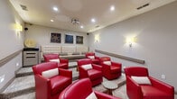 Located at 5003 Winster Dr in Winston-Salem, NC, this dedicated home theater room delivers an immersive cinematic experience within a thoughtfully designed single-level space. At the focal point of the room, a large projection screen anchors one wall, flanked by elegant drapes and complemented by a ceiling-mounted projector overhead. Recessed lighting and warm wall sconces cast a soft, ambient glow throughout, creating an inviting atmosphere ideal for movie viewing. The seating arrangement features multiple bold red leather club chairs accented with chevron-patterned throw pillows, arranged in tiered rows on a stylishly patterned carpet. A tufted cream-colored bench along the rear wall provides additional seating, while small accent tables are thoughtfully placed between chairs for convenience. Film reel artwork displayed on the back wall reinforces the cinematic theme. A handrail runs along the side wall to assist movement through the tiered layout. Surround sound speakers are mounted at strategic points throughout the room, ensuring high-quality audio. The neutral gray walls and carefully curated décor combine to create a polished, professional screening environment that balances comfort with style.