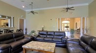 This single-story space offers a welcoming and well-appointed lounge area defined by its notably high ceilings adorned with crown molding and dual ceiling fans, contributing to an open and airy atmosphere. The seating area is anchored by a large dark leather sectional sofa and matching armchair, arranged around a stone-top coffee table with a wooden base, creating a comfortable and functional gathering space. Neutral beige walls provide a warm backdrop, complemented by decorative plants and a classic console table with a table lamp that adds a touch of warmth to the room. Toward the rear of the space, a striking columned archway frames a bright entryway, where an arched window above the entrance door floods the area with natural light, highlighting a decorative chandelier and potted plants that enhance the elegant aesthetic. The entryway's tiled flooring contrasts with the carpeted lounge area, further distinguishing the functional zones within the space. Overall, the design blends comfort with classic architectural details, making it an inviting environment suited for relaxation or social gathering.