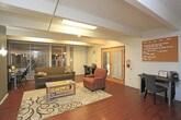 Located at 2201 Ambassador Rd NE in Albuquerque, New Mexico, this single-floor space is thoughtfully organized to serve as a functional community or resource center. The entrance greets visitors with a bold double-door entry flanked by wall sconces and dark accent walls, setting a grounded, purposeful tone. Rich hardwood flooring runs throughout, tying the various areas together cohesively. The main lounge area is furnished with a comfortable sofa, accent chairs, a coffee table, and a patterned area rug, creating an inviting gathering space. Large vertical-blind-covered windows look out toward an adjacent area, allowing natural light to filter in. A secondary workspace area features a round desk, office chair, and a cork bulletin board mounted on the wall, offering a focused setting for individual work or consultations. A fire extinguisher and window with horizontal blinds are also present in this area. Toward the back, a dedicated reading and resource room houses shelving units filled with books, a gear-shaped wall clock, wall art, and additional seating. HVAC venting is visible in the ceiling, ensuring year-round climate comfort. Overall, the space presents a practical, community-oriented environment well-suited for programming, drop-in services, or collaborative use.