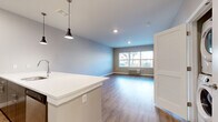 Located at 110 S Bridge St in Somerville, NJ, this thoughtfully designed one-bedroom, one-bathroom residence offers a clean, modern aesthetic throughout its well-utilized single floor. The open-concept kitchen and living area serves as the heart of the home, anchored by a large white quartz peninsula with an undermount sink and contemporary pendant lighting overhead. The kitchen is equipped with stainless steel appliances, including a refrigerator, microwave, and range, complemented by rich dark wood cabinetry and a classic subway tile backsplash. Wood-look plank flooring flows seamlessly throughout the entire space, adding warmth and visual continuity. The living area is bathed in natural light from a wide window that frames outdoor views of surrounding greenery. The spacious primary bedroom continues the light-filled theme with a large window, neutral wall tones, and ample room for furnishings. A dedicated closet provides convenient storage. The full bathroom features a dark wood vanity with a white countertop, a large mirror, and a subway-tiled soaking tub and shower combination. Clean lines, recessed lighting, and a neutral color palette give the entire residence a fresh, contemporary feel.