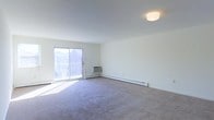This single-floor space features a generously sized, open living area finished with wall-to-wall carpet flooring and crisp white walls that create a clean, neutral backdrop. A sliding glass door along one wall fills the room with abundant natural light while providing direct access to an outdoor balcony, where a white railing is visible. The view through the door hints at a residential neighborhood setting. A wall-mounted air conditioning unit is positioned beneath the sliding door, complementing the baseboard heating that runs along the perimeter of the room for year-round climate control. The smooth, white ceiling is accented by a centrally placed flush-mount light fixture, contributing to the room's bright and airy atmosphere. The open layout offers a versatile living space well-suited for a variety of furniture arrangements. Overall, the space presents a straightforward, functional design characterized by ample natural light, practical climate control features, and a simple, uncluttered aesthetic.