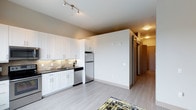 This single-floor space showcases a thoughtfully designed open-concept layout that seamlessly connects the kitchen and living areas. The kitchen is outfitted with white shaker-style cabinetry that extends to the ceiling, providing generous storage. A tasteful tile backsplash adds texture behind the dark countertops, which are complemented by a stainless steel sink with a modern faucet. Stainless steel appliances, including a refrigerator and dishwasher, complete the kitchen's contemporary look. Light gray luxury vinyl plank flooring runs throughout, lending a clean, cohesive feel to the space. A track lighting fixture on the smooth white ceiling provides focused illumination over the kitchen area, while a flush-mount ceiling light brightens the hallway beyond. A sliding barn-style door adds a functional yet stylish architectural detail, leading to what appears to be a closet or additional storage. The hallway connects to further areas of the unit, suggesting a practical and well-organized floor plan overall.