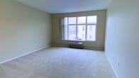 This single-floor space offers a clean, functional layout designed with simplicity and comfort in mind. The main living area is a standout feature, characterized by a large multi-pane window that floods the room with natural light while framing views of the neighboring urban architecture. The window spans a generous portion of the wall, creating an airy, open atmosphere that enhances the sense of space. Beneath the window sits a wall-mounted HVAC unit, providing convenient climate control directly within the living area. The room is finished with neutral beige carpeting that extends wall to wall, adding warmth and a soft underfoot feel. The walls are painted in a light, warm tone that complements the brightness brought in by the windows and contributes to the overall calm, understated ambiance of the space. Clean white baseboards provide a crisp contrast throughout. The layout is open and unobstructed, offering flexibility for a variety of furniture arrangements. The overall atmosphere is bright, quiet, and well-suited for both comfortable everyday living and urban convenience.