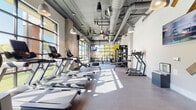This well-equipped fitness center in Charlotte's 28205 neighborhood offers a comprehensive, single-floor workout facility designed with both function and aesthetics in mind. The main cardio area features an impressive row of treadmills and other cardio machines set beneath soaring ceilings with exposed ductwork, pendant lighting, and oversized ceiling fans — giving the space an industrial-modern character. Large windows and glass garage-style doors flood the floor with abundant natural light, offering views of the surrounding outdoor areas and creating an open, energizing atmosphere. The strength training zone is stocked with a multi-functional cable machine system, free weights, benches, and specialty equipment, all positioned near floor-to-ceiling glass walls that connect visually to the outdoors. A dedicated group fitness and yoga studio features warm hardwood flooring, full-length mirrors, white brick walls, and glass partition doors that allow light to flow throughout. A cycling room with large grid-paned windows overlooking an outdoor terrace houses multiple stationary bikes with digital displays. Throughout the facility, the design balances industrial elements — including exposed ceilings and concrete finishes — with clean white walls and modern lighting, creating a motivating and visually cohesive environment for a full range of fitness activities.