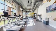 This well-equipped fitness center in Charlotte's 28205 neighborhood offers a comprehensive, single-floor workout facility designed with both function and aesthetics in mind. The main cardio area features an impressive row of treadmills and other cardio machines set beneath soaring ceilings with exposed ductwork, pendant lighting, and oversized ceiling fans — giving the space an industrial-modern character. Large windows and glass garage-style doors flood the floor with abundant natural light, offering views of the surrounding outdoor areas and creating an open, energizing atmosphere. The strength training zone is stocked with a multi-functional cable machine system, free weights, benches, and specialty equipment, all positioned near floor-to-ceiling glass walls that connect visually to the outdoors. A dedicated group fitness and yoga studio features warm hardwood flooring, full-length mirrors, white brick walls, and glass partition doors that allow light to flow throughout. A cycling room with large grid-paned windows overlooking an outdoor terrace houses multiple stationary bikes with digital displays. Throughout the facility, the design balances industrial elements — including exposed ceilings and concrete finishes — with clean white walls and modern lighting, creating a motivating and visually cohesive environment for a full range of fitness activities.