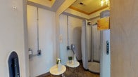 This single-floor tanning salon is designed to provide clients with a clean, functional, and inviting environment. The space is organized around a central corridor finished with large-format tile flooring in a cool gray tone, with warm wood trim framing the doorways throughout. The hallway connects multiple private tanning rooms and leads toward a naturally lit seating area at the far end, furnished with upholstered chairs and a small table.

Each tanning room is equipped with a stand-up tanning unit featuring wraparound UV panels and a private entry door, clearly marked with operational signage. The rooms include small accent tables with decorative greenery and sanitation supplies, maintaining a hygienic and organized atmosphere. Electrical panels and control units are neatly mounted on the walls, and large mirrors enhance the sense of space within each room. The flooring transitions to a darker stone-look tile within the tanning areas, providing visual contrast.

Decorative touches, including a sculptural sun wall accent and tufted seating in one of the prep areas, add warmth and reinforce the salon's sun-themed aesthetic. The overall ambiance is clean, professional, and welcoming, with practical design elements supporting efficient client flow.