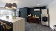 The Hydration Room is a single-floor amenity space designed to serve as a convenient refreshment hub. The room features a clean, functional layout anchored by a light wood-toned central island with seating, providing a casual gathering point for occupants. Textured carpet flooring in a dark, multi-toned pattern runs throughout, complemented by neutral-toned walls and a textured ceiling with recessed lighting.

A bold navy accent wall serves as a focal point, housing a dark wood beverage and coffee station with granite-style countertops, alongside a wall-mounted mini-split HVAC unit. Adjacent to this area, a dedicated vending alcove features both a beverage and a snack machine, positioned near large windows that allow natural light to fill the space. An ice machine rounds out the beverage offerings nearby.

Interior sliding windows connect the room to an adjacent area, enhancing visibility and accessibility. A gray panel entry door with a classic six-panel design provides secure access. The overall ambiance is practical yet welcoming, balancing commercial-grade amenities with a tidy, modern aesthetic suited for a communal refreshment area.