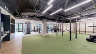 Located at 49 Fisk St in Jersey City, NJ, this professionally designed fitness center offers a generously sized, single-floor layout accommodating a full range of workout zones. The main gym area features an industrial-modern aesthetic, with an exposed ceiling showcasing dark-painted ductwork and structural elements alongside sleek linear LED lighting that illuminates the space efficiently. Large windows along the perimeter allow natural light to flow in while offering views of the surrounding outdoors. The gym is equipped with cardio machines including treadmills and ellipticals, a variety of strength training cable machines, and dedicated free weight stations. A expansive turf-covered functional training zone anchors the center of the space, outfitted with power racks, kettlebells, medicine balls, battle ropes, and other functional fitness accessories. Built-in shelving units provide organized storage for equipment. A separate studio room features warm hardwood flooring, a tray ceiling with recessed linear lighting, wall-mounted display screens, and a clean, minimalist design ideal for group fitness classes, yoga, or stretching. Exposed concrete columns throughout the main area add an urban, contemporary character that ties the entire facility together into a cohesive and high-quality fitness environment.
