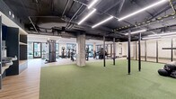 Located at 49 Fisk St in Jersey City, NJ, this professionally designed fitness center offers a generously sized, single-floor layout accommodating a full range of workout zones. The main gym area features an industrial-modern aesthetic, with an exposed ceiling showcasing dark-painted ductwork and structural elements alongside sleek linear LED lighting that illuminates the space efficiently. Large windows along the perimeter allow natural light to flow in while offering views of the surrounding outdoors. The gym is equipped with cardio machines including treadmills and ellipticals, a variety of strength training cable machines, and dedicated free weight stations. A expansive turf-covered functional training zone anchors the center of the space, outfitted with power racks, kettlebells, medicine balls, battle ropes, and other functional fitness accessories. Built-in shelving units provide organized storage for equipment. A separate studio room features warm hardwood flooring, a tray ceiling with recessed linear lighting, wall-mounted display screens, and a clean, minimalist design ideal for group fitness classes, yoga, or stretching. Exposed concrete columns throughout the main area add an urban, contemporary character that ties the entire facility together into a cohesive and high-quality fitness environment.