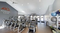 This fitness center offers a comprehensive cardio-focused workout environment designed to meet a range of exercise needs. The space is outfitted with multiple elliptical machines, stair climbers, and treadmills, arranged to maximize accessibility and flow throughout the room. The vaulted ceiling enhances the sense of openness, while recessed lighting and a ceiling fan provide a comfortable, well-lit atmosphere. Natural light filters in through several windows along the walls, adding brightness and an inviting feel to the space. The flooring transitions between a rubberized mat area beneath the cardio equipment and a smooth dark surface, providing both functionality and durability. The walls are finished in a neutral gray tone, contributing to a clean, modern aesthetic. A dumbbell rack is visible along one wall, offering supplementary free weight options. Overall, the fitness center combines practical equipment layout with a well-maintained, contemporary design to support an effective and comfortable exercise experience.