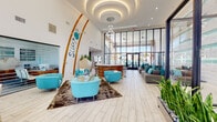 Surf2 is a thoughtfully designed community amenity space that embraces a bold coastal aesthetic from the moment you step inside. The grand lobby immediately captures attention with soaring ceilings, dramatic circular layered cove lighting, and a cascading ring chandelier. Decorative oversized surfboards flank the feature wall bearing the Surf2 logo, while turquoise lounge chairs, a glass-top coral coffee table, and a circular reception desk set a lively, beach-inspired tone. Light wood-look tile flooring and floor-to-ceiling glass walls flood the space with natural light and outdoor views.

Adjacent to the lobby, a relaxing lounge area features a sleek linear fireplace set against a sculptural white 3D-textured accent wall with teal LED shelf lighting, surrounded by plush grey sofas and teal accent pillows. A casual bar nook with high-top tables and a wine refrigerator offers a social retreat with pool views.

The glass-enclosed e-lounge provides a bright, functional workspace, while the fitness wing delivers a comprehensive wellness experience. The weight room offers a full dumbbell rack, cable machines, and mirrored walls, while a dedicated studio space with spin bikes, TRX suspension trainers, wall balls, and hardwood flooring supports group and individual training. Teal accent walls and ceiling fans are consistent throughout, tying the entire space together with energetic, resort-style character.
