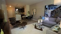 This well-appointed single-floor residence showcases a thoughtfully designed open-concept layout that seamlessly connects the kitchen, dining, and living areas into one cohesive, functional space. The kitchen features dark cabinetry with modern hardware, stainless steel appliances, and a light-toned countertop that contrasts elegantly with the darker finishes. Pendant lighting hangs above, adding a warm, focused glow to the culinary space. Adjacent to the kitchen, the dining area accommodates a round table with sleek black chairs, making it an intimate spot for everyday meals or entertaining guests. The living area is anchored by a plush white area rug and a distinctive glass-top coffee table with a sculptural wooden base, complemented by a classic leather lounge chair that adds a timeless touch. Neutral warm-gray walls provide a versatile backdrop, accented by curated artwork that adds personality and color to the space. Light wood-tone flooring flows throughout, tying the rooms together and enhancing the sense of openness. The overall ambiance is modern yet inviting, balancing clean lines with warm textures for a comfortable, contemporary feel.