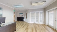 Located on Franklin Street in Hampton, Virginia, this single-floor facility is thoughtfully designed to serve as a welcoming and functional community or leasing office space. Light wood-tone luxury vinyl plank flooring flows consistently throughout, tying the various areas together with a clean, cohesive aesthetic.

The entry area features elegant French doors with shuttered glass panels that invite natural light into the space, creating a bright and open atmosphere. Crown molding adds a refined architectural touch throughout. A dedicated reception area includes a substantial front desk, coastal-inspired artwork, and a comfortable seating chair, establishing a professional yet inviting first impression.

Adjacent to the entry is a spacious lounge and sitting area, highlighted by a bold teal accent wall that provides a vibrant focal point. Comfortable seating arrangements and abundant natural light from multiple windows make this area ideal for relaxation or informal gatherings. A separate dining area, also accented with a deep teal wall, accommodates multiple round glass-top tables beneath a modern multi-globe chandelier.

A private office with large windows, navy drapery, and a built-in workstation offers a quiet workspace. The fully equipped kitchen features warm wood cabinetry, granite countertops, a stainless steel appliance suite, and ample storage, providing practical amenities for staff and residents alike.