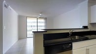 This single-floor unit showcases an open-concept layout that seamlessly connects the kitchen and living areas, creating a functional and airy living environment. The kitchen is outfitted with dark countertops, a built-in dishwasher, and a sink with modern fixtures. A peninsula-style counter serves as a natural divider between the kitchen and the main living space, offering additional workspace and a casual seating opportunity. Light-toned tile flooring runs throughout, contributing to a clean, cohesive aesthetic. A ceiling fan is centrally positioned overhead, providing practical climate control. The living area is anchored by expansive floor-to-ceiling sliding glass windows and doors that flood the space with abundant natural light. These open onto a private balcony, offering views of the surrounding urban exterior. The white walls and textured ceiling keep the interior feeling bright and neutral, providing a versatile backdrop for a range of interior styles. Overall, the space delivers a practical and well-lit open-plan design with the added benefit of outdoor access and generous daylighting throughout.