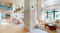 Located at 2480 First St in Fort Myers, Florida, this sophisticated two-story space offers a seamlessly flowing interior defined by refined coastal design and premium materials. The expansive main floor is anchored by dramatic double-height ceilings that flood the space with natural light through floor-to-ceiling black-framed windows, framing views of swaying palm trees and lush Florida greenery. Distinctive travertine-clad columns rise through the open plan, complemented by warm wide-plank hardwood flooring and tongue-and-groove wood ceiling panels that lend a rich, organic warmth throughout.

The main floor encompasses multiple distinct zones, including a generous lounge area with a pool table, a sunlit seating nook with a plush wraparound sofa, and a long communal bar-height table beneath sculptural woven pendant lights. Small-group seating areas with curved furnishings and built-in banquettes provide flexible gathering spaces. A dedicated café-style section features compact round tables and custom millwork shelving.

The upper level, accessed via a striking staircase with glass-panel railings, offers a more intimate atmosphere with built-in lounge alcoves, warm brass wall sconces, and open-railed mezzanine views overlooking the main floor below. Bright windows along the upper level continue the connection to the outdoors, making this a light-filled and thoughtfully designed destination throughout.