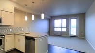 This single-floor space showcases a thoughtfully designed open-concept layout that seamlessly connects the kitchen and living area. The kitchen is a standout feature, centered around a peninsula island with a sleek granite-style countertop, an undermount sink with a modern arc faucet, and a stainless steel dishwasher. Three elegant cylindrical pendant lights hang above the island, providing both functional task lighting and a stylish focal point. A subway tile backsplash adds a classic yet contemporary touch to the kitchen wall. The living area flows naturally from the kitchen, finished with light-toned carpet that contrasts beautifully with the dark hardwood flooring in the kitchen zone. Large windows along the far wall invite an abundance of natural light, creating a bright and airy ambiance throughout the space. A glass door provides additional access and further enhances the sense of openness. The neutral gray wall tones throughout lend a clean, modern aesthetic that complements the overall design. The space is practical yet refined, offering a comfortable and well-lit environment well-suited for everyday living.