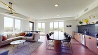 This stylish, single-floor space offers an open-plan layout that seamlessly integrates multiple functional zones, creating a versatile environment suited for both collaborative gatherings and relaxed socializing. Rich hardwood flooring flows throughout, unifying the space with warmth and continuity.

The kitchen is a standout feature, boasting a substantial curved island topped with a dark stone countertop and barstool seating. Warm wood cabinetry with gold hardware pairs elegantly with a decorative mosaic tile backsplash, illuminated by brass wall sconces. A stainless steel refrigerator and full suite of appliances complete the well-equipped setup.

Adjacent to the kitchen, a large oval conference-style dining table surrounded by sleek black chairs provides ample seating for group meetings or communal dining. This area transitions naturally into a generously sized lounge, where a sectional sofa, mid-century accent chairs, and a round coffee table are arranged around a plush area rug. A wall-mounted flat-screen TV anchors one end of the lounge, while a striking gold ceiling fan adds a designer touch overhead.

Large floor-to-ceiling windows on multiple walls flood the space with abundant natural light and frame expansive outdoor views of the surrounding neighborhood and greenery, creating a bright and airy ambiance throughout.