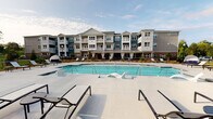 This outdoor amenity space is designed to offer a resort-like experience, centered around a generously sized swimming pool with crystal-clear blue water. The pool deck is finished in light-toned concrete and furnished with a variety of chaise lounges and accent tables, providing ample space for sunbathing and relaxation. Unique in-pool lounge chairs allow residents to recline directly in the water, enhancing the leisure-focused design. The pool area is framed by a sleek iron perimeter fence, with lush landscaping adding a natural backdrop.

Adjacent to the pool, a covered outdoor lounge area serves as a shaded gathering space. This pavilion features a bold teal, paneled accent wall that anchors the space visually and is equipped with a wall-mounted flat-screen television and wall sconces. The seating arrangement includes wicker armchairs and a sofa set around a coffee table, complemented by additional dining-height seating nearby. A built-in outdoor grill station is also present, making the space well-suited for entertaining. The cobblestone-style patio flooring adds texture and character throughout. The backdrop includes a multi-story residential building with blue and gray exterior siding and private balconies, completing the cohesive community setting.